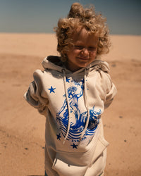 Kind mit blonden Locken steht am Strand im Ankerherz Hoodie EUROPA ONE LOVE Ivory Garment Dye, blauer Siebdruck Europa auf Zeus-Stier mit zwölf Sternen von Dani Garreton, Unisex Frontansicht