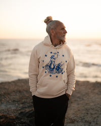 Mann steht am Strand im Ankerherz Hoodie EUROPA ONE LOVE Ivory Garment Dye, blauer Siebdruck Europa auf Zeus-Stier mit zwölf Sternen von Dani Garreton, Unisex Frontansicht