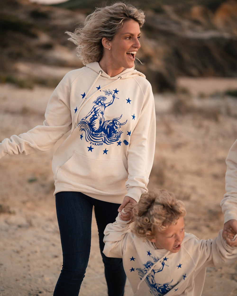 Frau und Kind toben am Strand im Ankerherz Hoodie EUROPA ONE LOVE Ivory Garment Dye, blauer Siebdruck Europa auf Zeus-Stier mit zwölf Sternen von Dani Garreton, Unisex Frontansicht