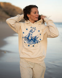 Frau läuft über den Strand im Ankerherz Hoodie EUROPA ONE LOVE Ivory Garment Dye, blauer Siebdruck Europa auf Zeus-Stier mit zwölf Sternen von Dani Garreton, Unisex Frontansicht